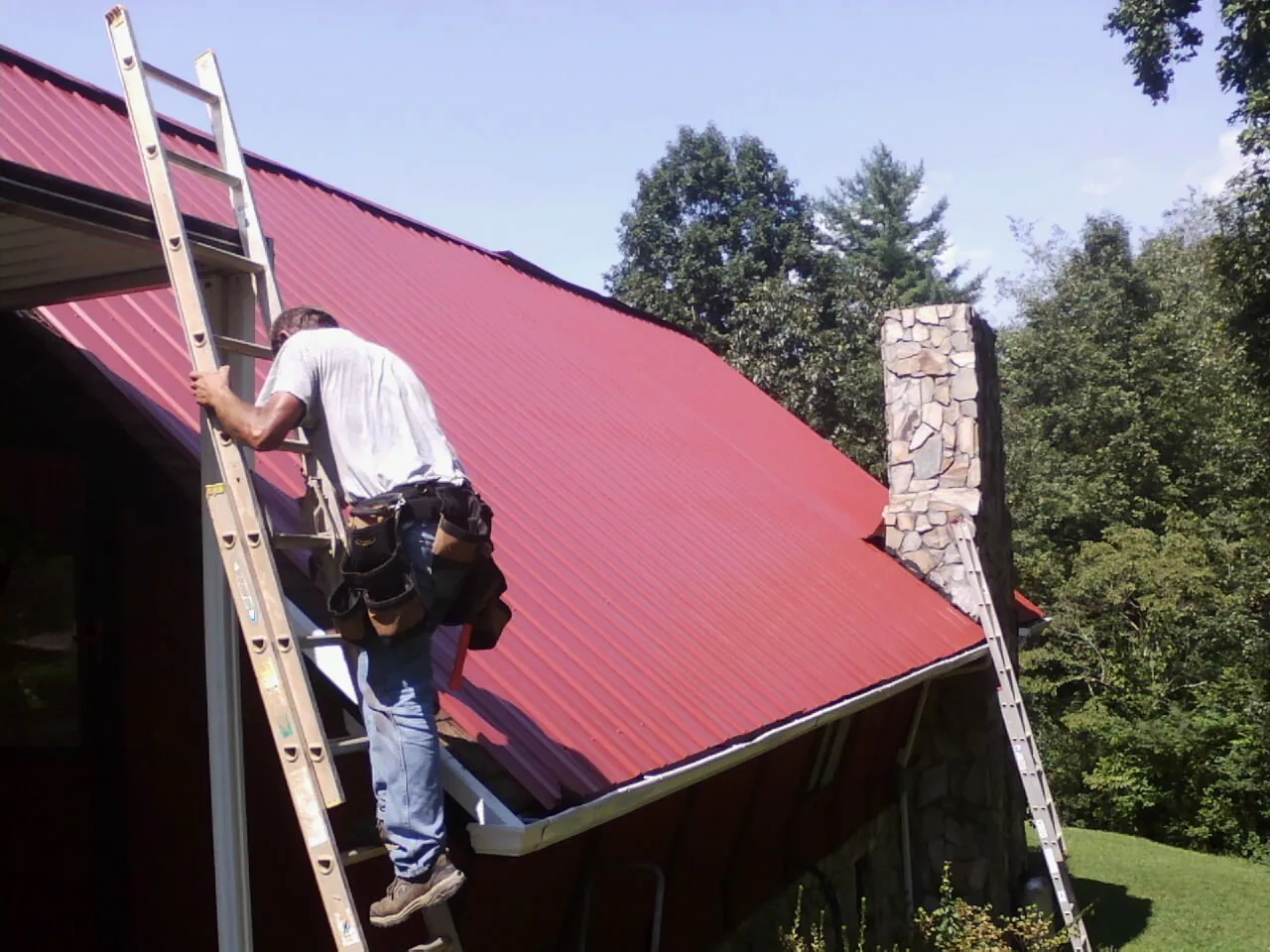 Roofer on a ladder beside a red metal roof and stone chimney — Soffit & Fascia Repair by Roofing Brothers in Larue